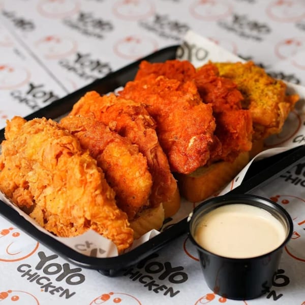 A black tray of Yoyo Chicken's fried chicken tenders, some spicy red, with a side of dipping sauce.