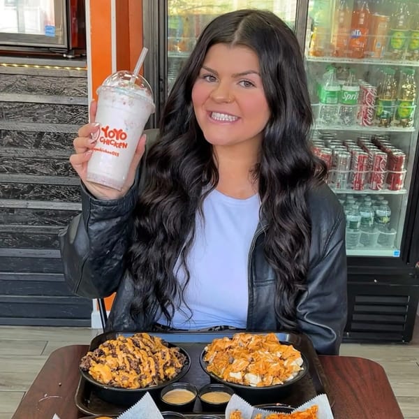 A smiling woman holds up a Yoyo Chicken milkshake, with fried chicken and loaded fries on the table.