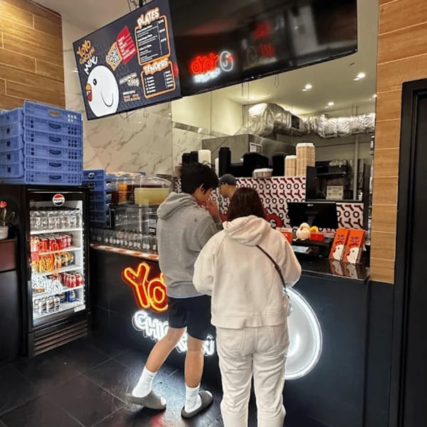 Two customers stand at the counter of Yoyo Chicken, with a menu board and drink fridge visible.