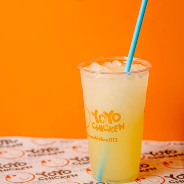 Yoyo Chicken branded lemonade with ice and a blue straw on a patterned paper, against an orange background.